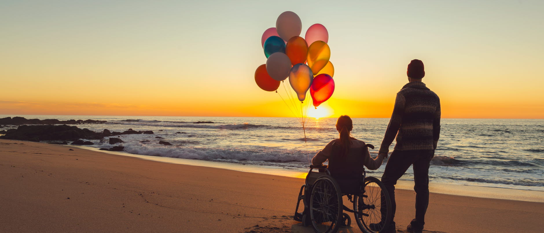 Två personer står på en strand med solnedgång som går ner över vattnet. Person i rullstol håller i ballonger.